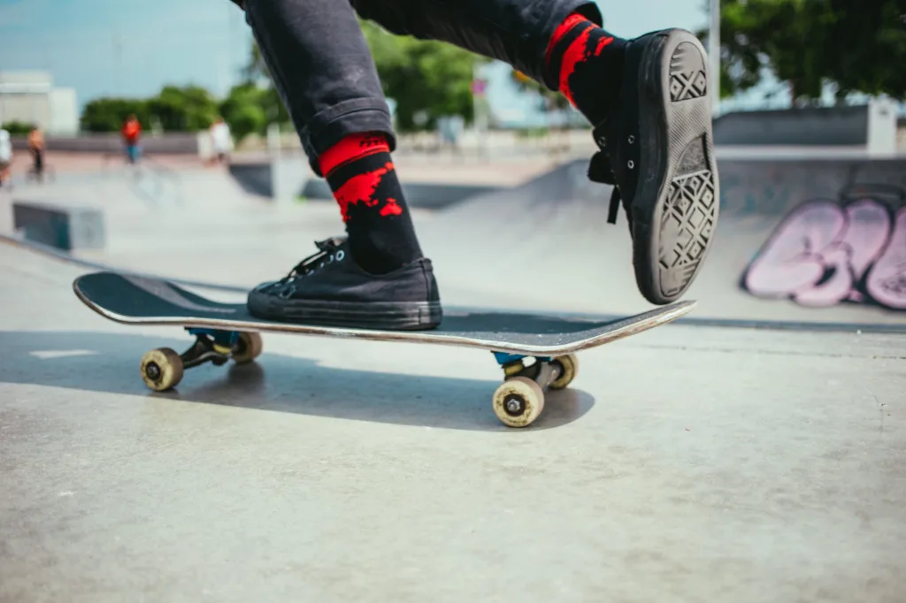 Close-up of a person riding a skateboard, focusing on their feet and the board. One foot is on the board, while the other foot is up, in motion. A skatepark is blurred in the background.